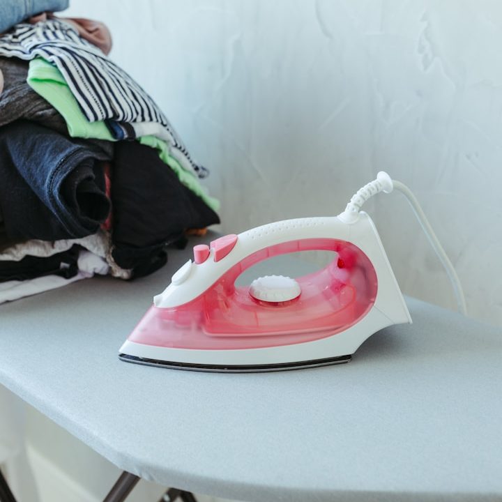 A pink and white steam iron on an ironing board beside stacked clothes.