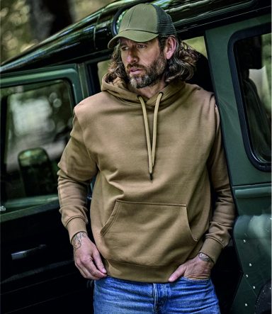 Man in a brown hoodie and cap leans against a vehicle in a forested setting.