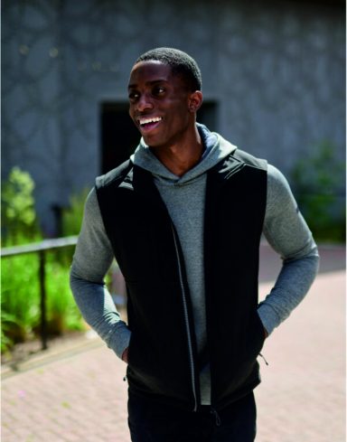 Smiling man in a grey hoodie and black vest standing outdoors.