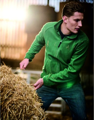 Man in a green jacket stacking hay in a barn with warm lighting.